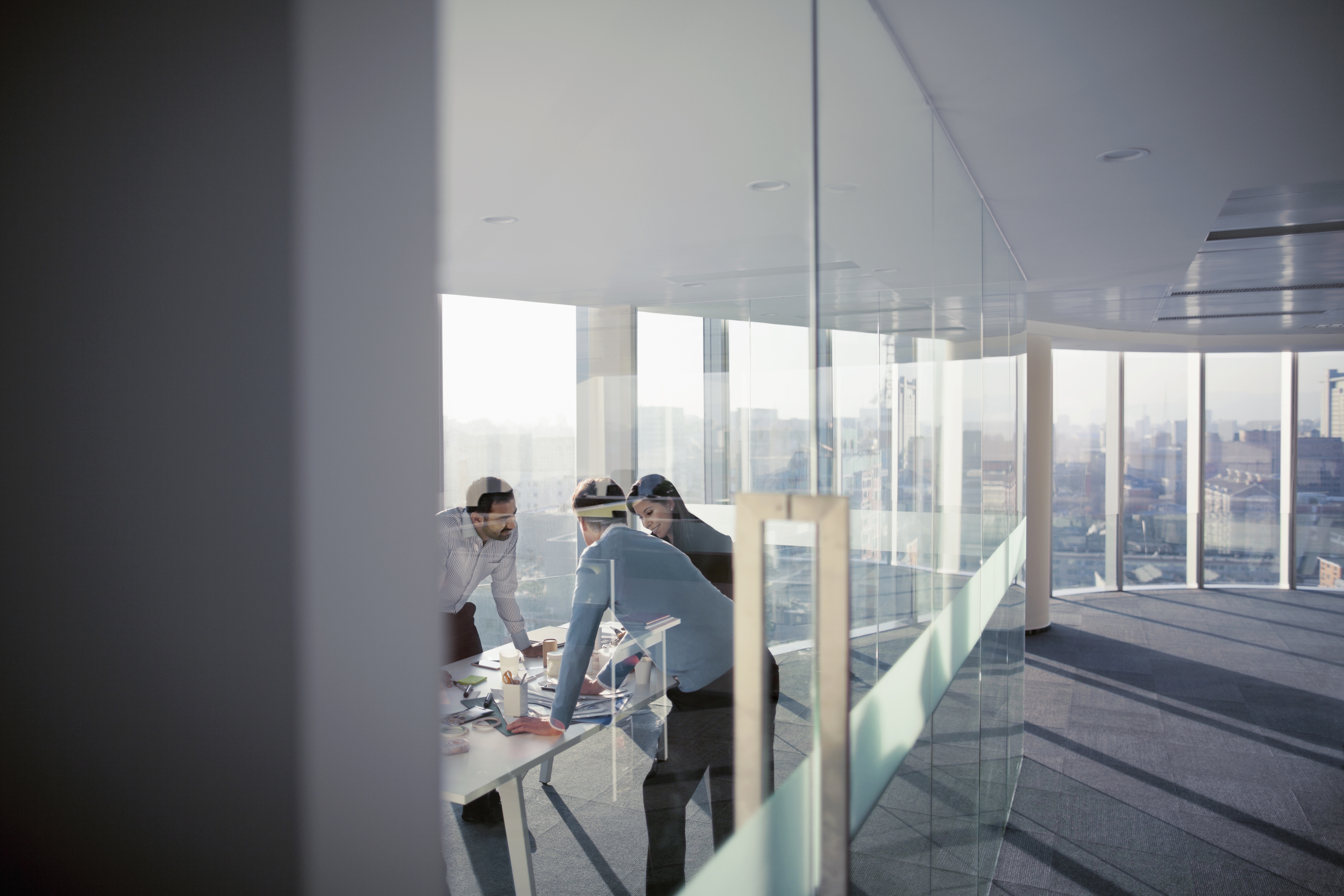 three team members leaning over a table in an office surrounded by glass walls
